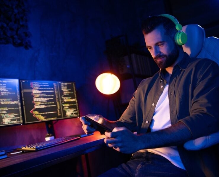 A man writing code, looking at his phone with headphones on